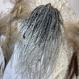 May include: A set of gray and white dreadlocks with a textured appearance. The dreadlocks are arranged in a bundle, with some strands cascading downwards. The image includes decorative elements like dried plants and a lace doily.