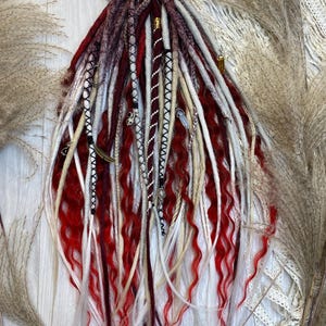 Könnte beinhalten: Eine Sammlung roter, weißer und burgunderfarbener Dreadlocks, verziert mit dekorativen Perlen und Charms. Die Dreadlocks haben eine wellige Textur und sind vor dem Hintergrund von getrocknetem Ziergras und Spitze angeordnet.