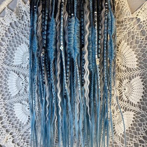 May include: A collection of blue, gray, and white dreadlocks hanging from a gold hanger. The dreadlocks vary in texture, with some being smooth and others wavy. Some are adorned with small beads. The background is a white, lacy dreamcatcher.