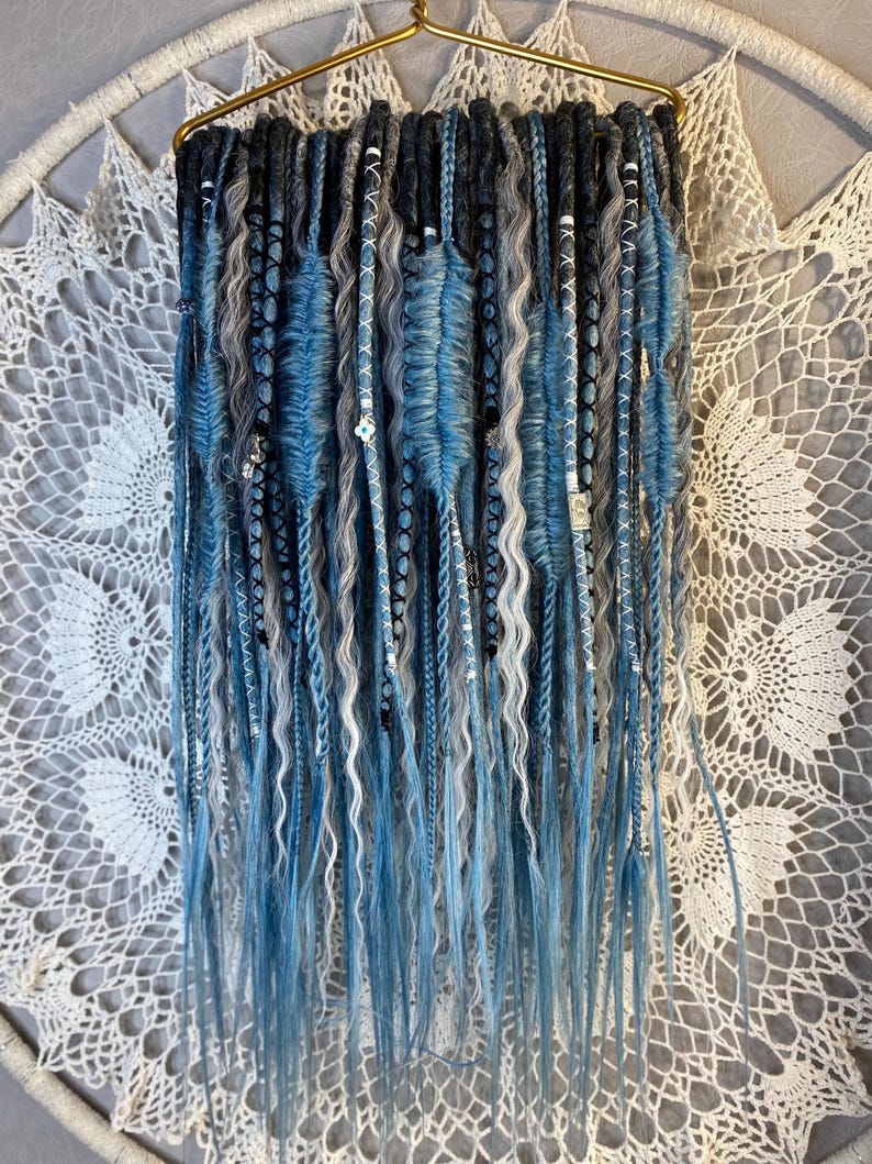 May include: A collection of blue, gray, and white dreadlocks hanging from a gold hanger. The dreadlocks vary in texture, with some being smooth and others wavy. Some are adorned with small beads. The background is a white, lacy dreamcatcher.