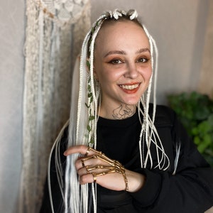 May include: A gold-colored skeleton hand bracelet with matching rings. The person has white braided hair with green leaves and a black top. A dreamcatcher hangs in the background.
