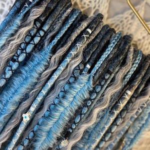 May include: A collection of blue, gray, and black dreadlocks, some with braided sections and white thread accents. The dreadlocks are arranged on a gold hanger, showcasing a variety of textures and colors.