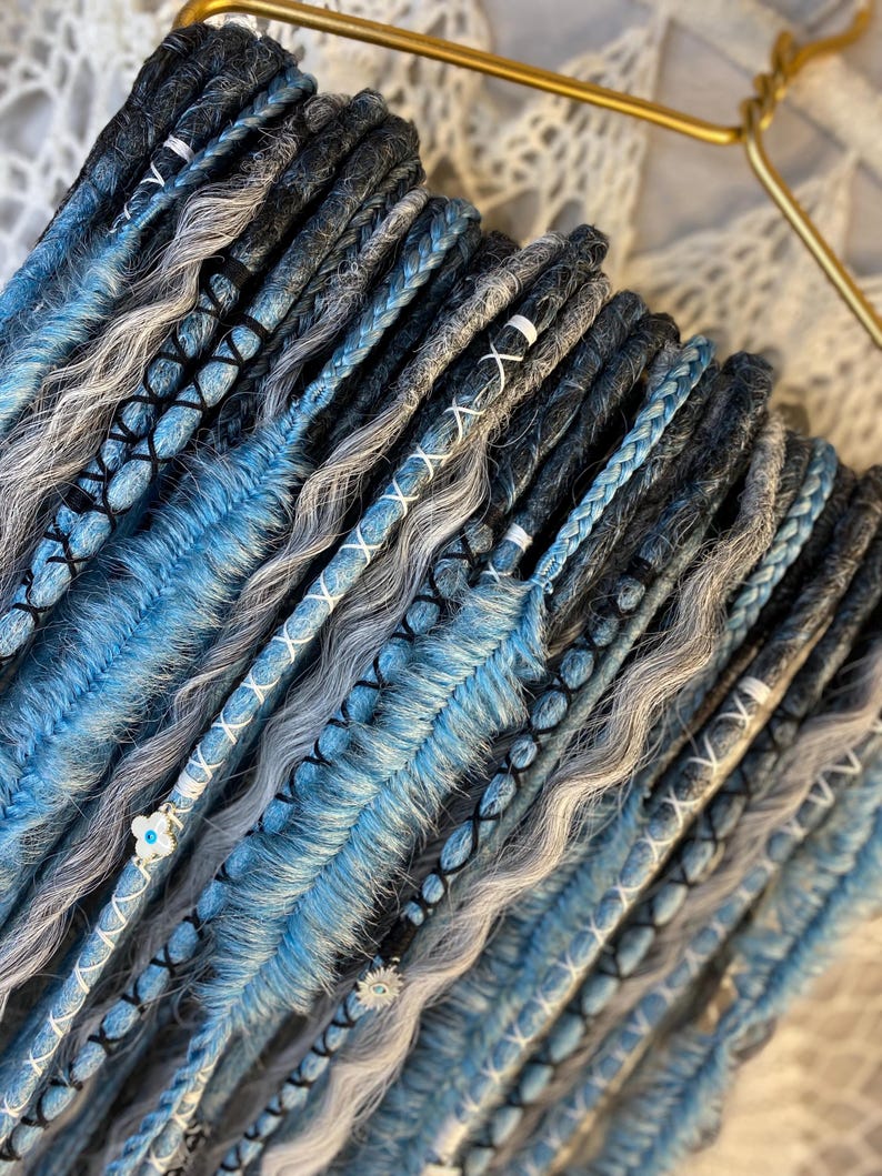 May include: A collection of blue, gray, and black dreadlocks, some with braided sections and white thread accents. The dreadlocks are arranged on a gold hanger, showcasing a variety of textures and colors.