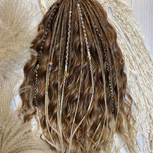 May include: A set of brown and blonde dreadlocks with wavy ends. The dreadlocks are adorned with decorative beads and white and black string. The hair is styled on a white surface with a cream-colored lace doily and dried ornamental grass.