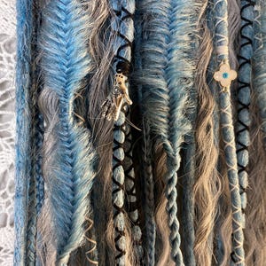 May include: Close-up of handmade dreadlocks in shades of blue, gray, and black. The dreads feature intricate braiding, wrapped sections, and decorative charms. The overall aesthetic is bohemian and artistic.