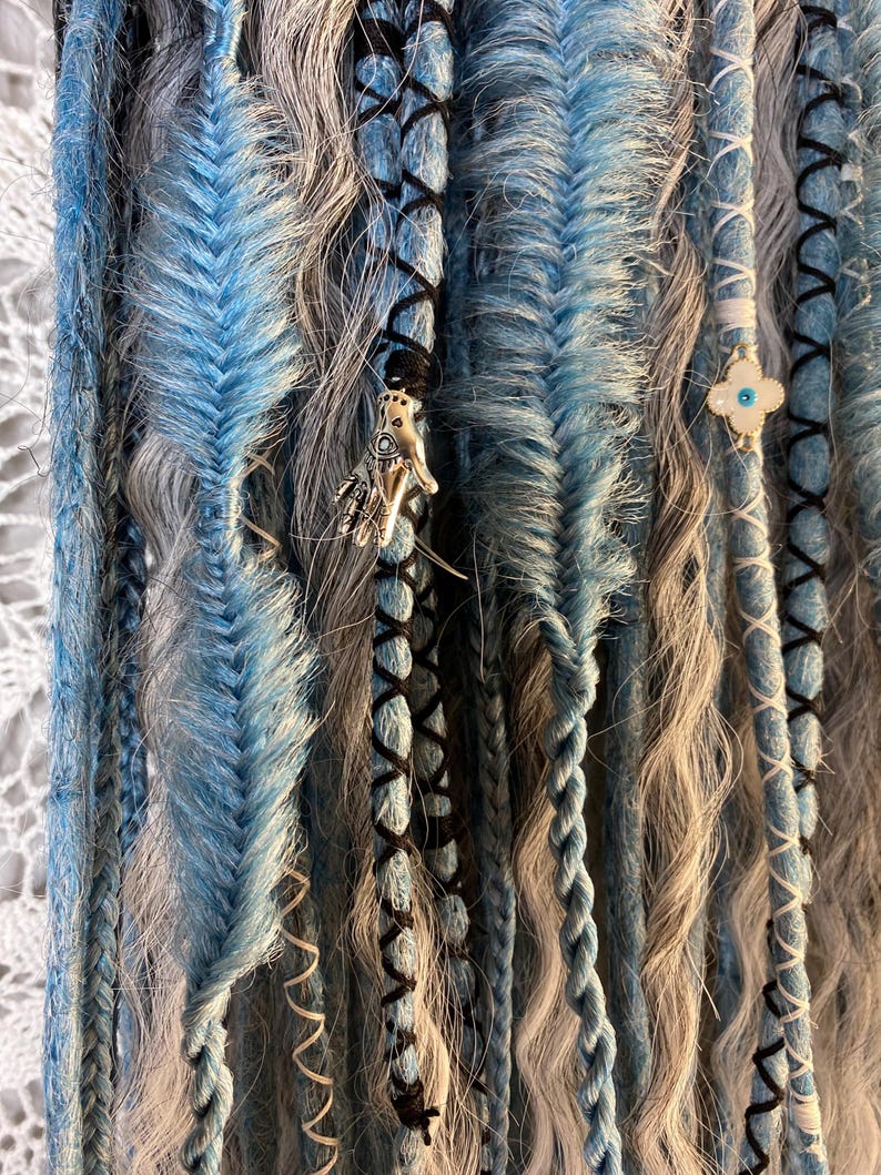 May include: Close-up of handmade dreadlocks in shades of blue, gray, and black. The dreads feature intricate braiding, wrapped sections, and decorative charms. The overall aesthetic is bohemian and artistic.