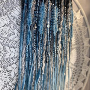 May include: A collection of blue, gray, and white dreadlocks, some with decorative accents. The dreadlocks are arranged in a vertical display, with varying textures and shades of blue. The background features a white, lacy textile.
