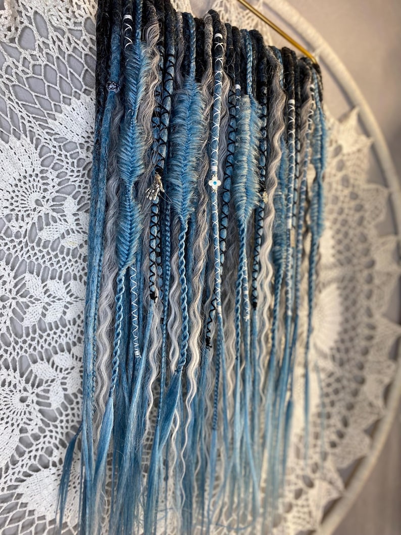 May include: A collection of blue, gray, and white dreadlocks, some with decorative accents. The dreadlocks are arranged in a vertical display, with varying textures and shades of blue. The background features a white, lacy textile.