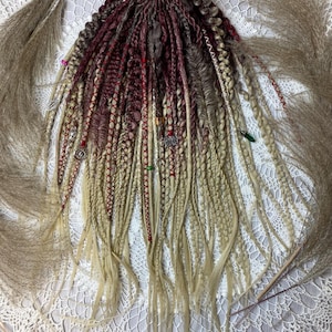 May include: A set of synthetic hair extensions featuring a blend of deep burgundy, brown, and blonde strands. The hair is styled into various textures including tight braids, twists, and loose waves, accented with small silver charms and beads.