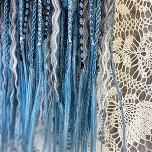 May include: Close-up of blue, white, and gray dreadlocks. The dreads are styled with various patterns, including braids and wrapped sections. Some dreads have white beads and black accents. The background is a white, lacy fabric.