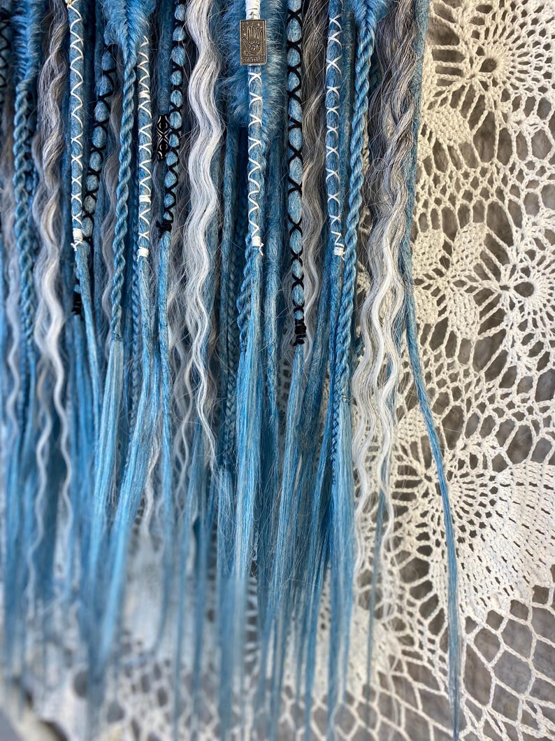 May include: Close-up of blue, white, and gray dreadlocks. The dreads are styled with various patterns, including braids and wrapped sections. Some dreads have white beads and black accents. The background is a white, lacy fabric.