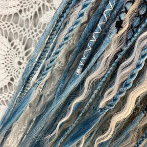 May include: Close-up of blue, white, and gray dreadlocks. The dreadlocks are a mix of braided and loose strands, with some sections wrapped in white thread. The color palette includes shades of blue, white, and gray, creating a textured and layered appearance.