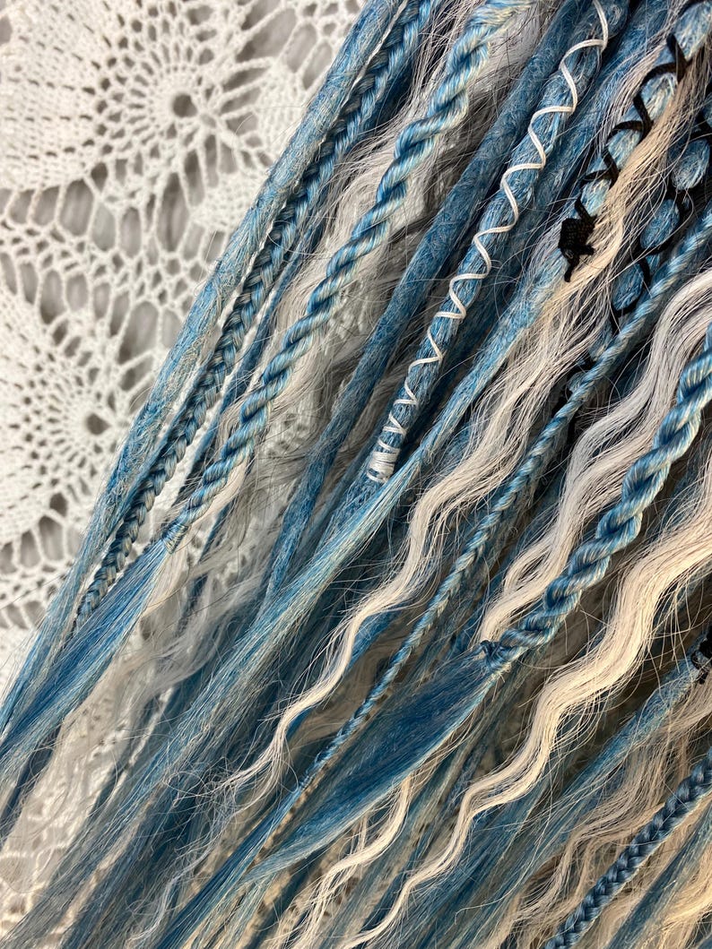 May include: Close-up of blue, white, and gray dreadlocks. The dreadlocks are a mix of braided and loose strands, with some sections wrapped in white thread. The color palette includes shades of blue, white, and gray, creating a textured and layered appearance.