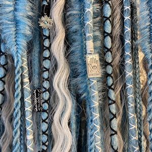 May include: Close-up of blue, gray, and white dreadlocks with decorative charms. The dreadlocks are adorned with black and white stitching and small metal charms, including an eye and a charm with the word "FORTUNE".
