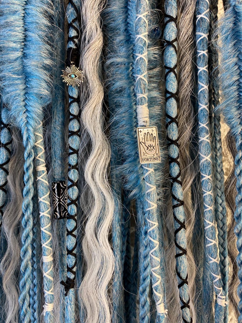 May include: Close-up of blue, gray, and white dreadlocks with decorative charms. The dreadlocks are adorned with black and white stitching and small metal charms, including an eye and a charm with the word "FORTUNE".