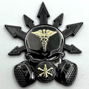 May include: Black and gold skull pin with a medical caduceus symbol on the forehead and a gas mask design. The skull is surrounded by eight black points radiating outward.
