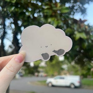 May include: A white cloud-shaped sticker with a smiling face and two small, dark gray wheels. The sticker is held up against a blurred background of green foliage, a street, and a white vehicle.