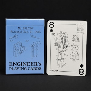 May include: A light blue deck of Engineer's Playing Cards box with patent text and diagrams, next to a white playing card featuring a black line drawing of a space suit labeled Fig. 5 and Fig. 11 with the number 8 in the corners.
