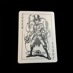 May include: A playing card featuring a steampunk-style Joker illustration in black and white. The Joker is depicted with gears and mechanical elements, wearing a top hat and holding a gear. The word "JOKER" is printed vertically on the card.