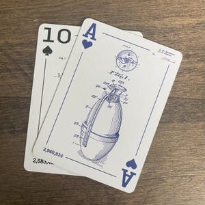 Two playing cards on a wooden surface. The top card is the Ace of Spades, featuring an engineering drawing of a grenade with the text 'FIG. 1.' and patent information. The bottom card is the Ten of Spades. Both cards have a white background with black suit symbols and text.