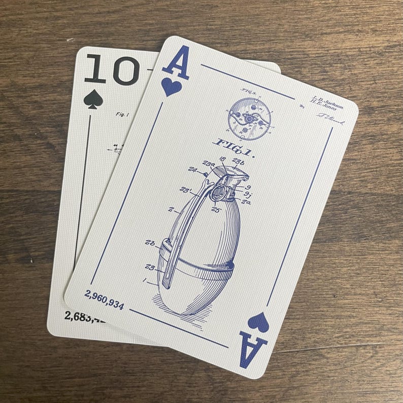 Two playing cards on a wooden surface. The top card is the Ace of Spades, featuring an engineering drawing of a grenade with the text 'FIG. 1.' and patent information. The bottom card is the Ten of Spades. Both cards have a white background with black suit symbols and text.