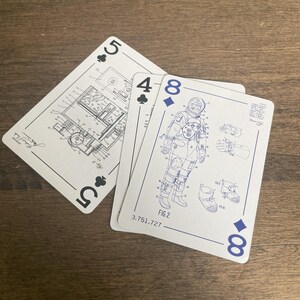 May include: Three playing cards are displayed on a wooden surface. The cards are white with blue ink designs. One card shows a five of clubs with a lighter technical drawing. Another card shows a four of clubs with a covered technical drawing. The third card shows an eight of diamonds with a technical drawing of a NASA astronaut.