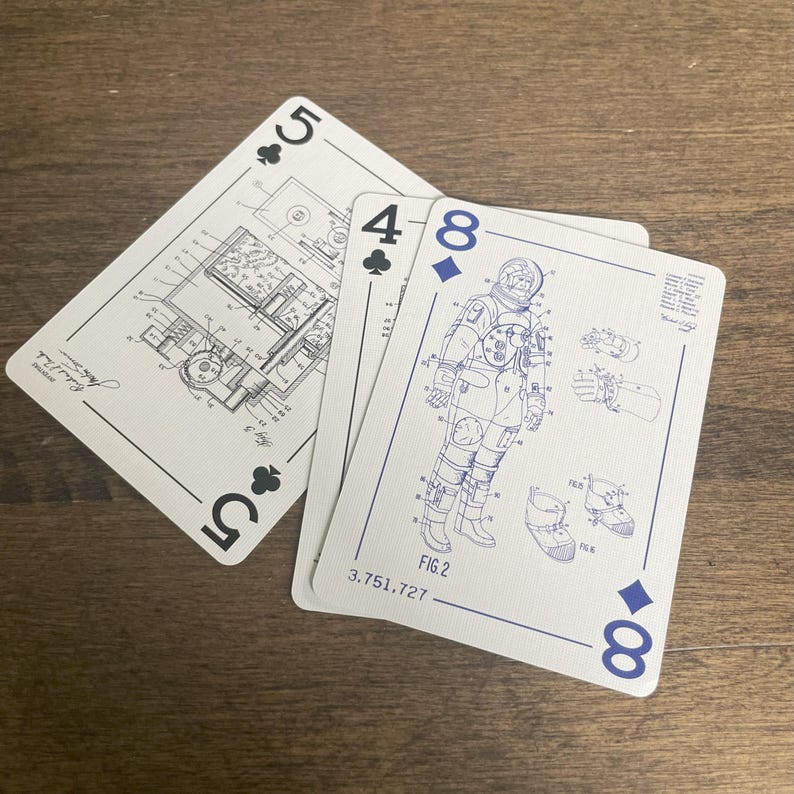May include: Three playing cards are displayed on a wooden surface. The cards are white with blue ink designs. One card shows a five of clubs with a lighter technical drawing. Another card shows a four of clubs with a covered technical drawing. The third card shows an eight of diamonds with a technical drawing of a NASA astronaut.