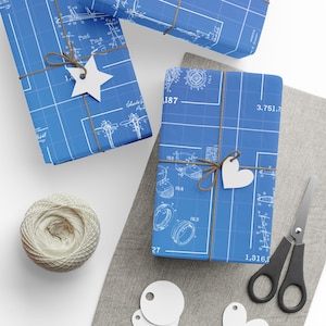 May include: Blue gift wrap with a blueprint design, tied with twine and adorned with white star and heart-shaped tags. A ball of twine, scissors, and additional tags are also present, suggesting a DIY gift-wrapping theme.
