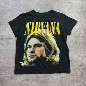 Puede incluir: Camiseta negra con el nombre de la banda "NIRVANA" en letras amarillas sobre un retrato de un hombre con pelo largo y rubio. La camiseta de manga corta está hecha de un material suave.