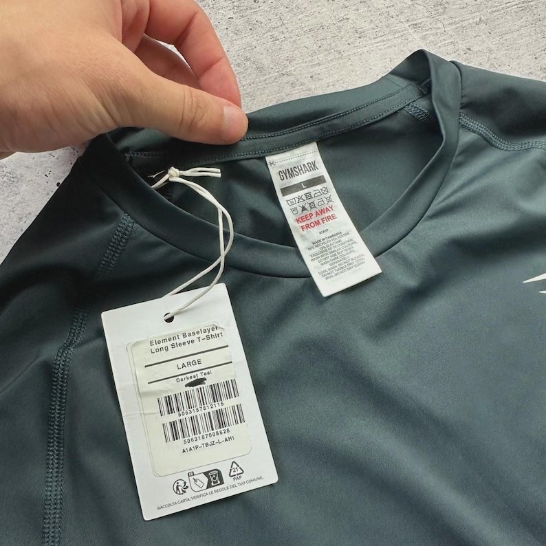 May include: Dark teal long-sleeve Element Baselayer T-shirt, size large. The shirt has a tag that reads "Gymshark" and "Keep Away From Fire". The shirt is a solid color with a tag attached.