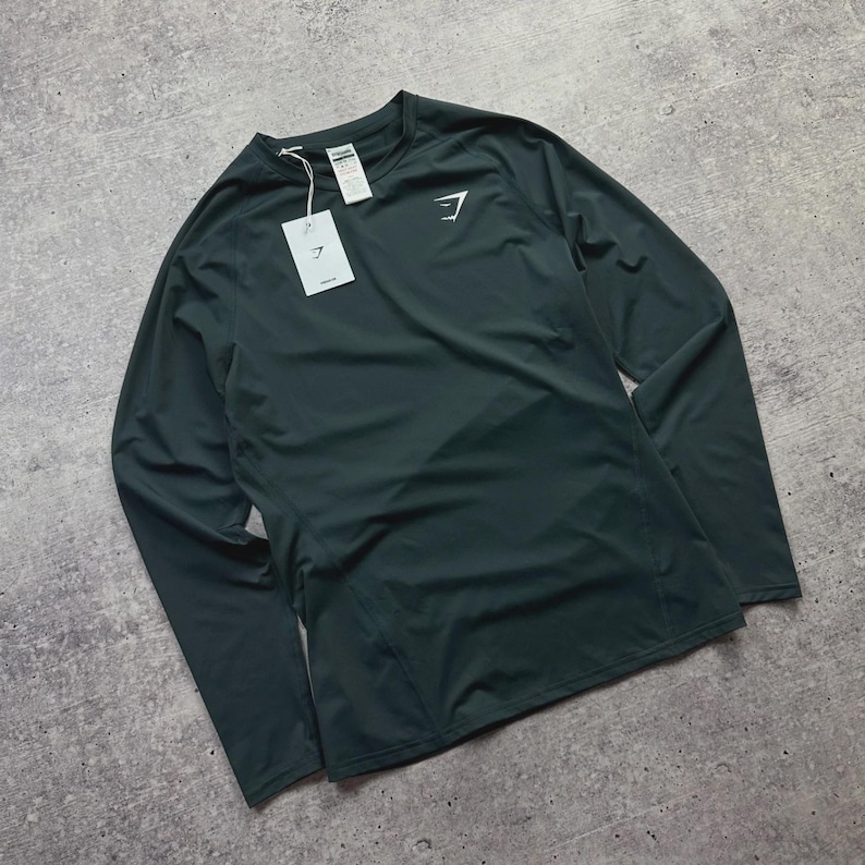 May include: Dark green long-sleeve athletic shirt with a crew neck. The shirt has a white logo on the chest and a tag attached to the collar. The shirt is made of a lightweight, breathable material.