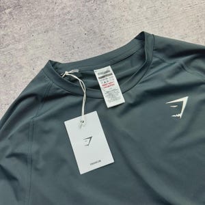 May include: A teal-colored athletic shirt with a crew neck and raglan sleeves. The shirt features a white Gymshark logo on the chest and a tag with the Gymshark brand name and care instructions. The shirt is made of a lightweight, breathable fabric.