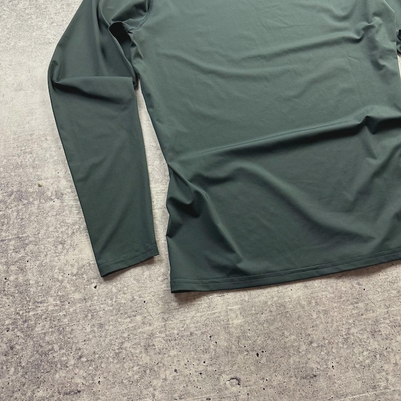 May include: A long-sleeved, dark green shirt. The shirt is made of a smooth, possibly athletic material. The sleeves are long and the shirt appears to be a close fit. The background is a textured, gray surface.