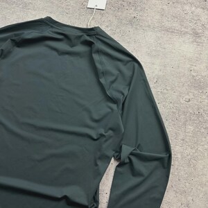 May include: A dark teal long-sleeve shirt with a crew neck. The shirt is made of a smooth, possibly athletic material. A white tag hangs from the neckline.