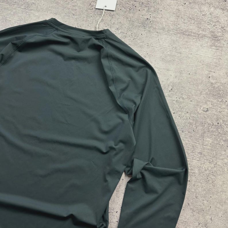 May include: A dark teal long-sleeve shirt with a crew neck. The shirt is made of a smooth, possibly athletic material. A white tag hangs from the neckline.