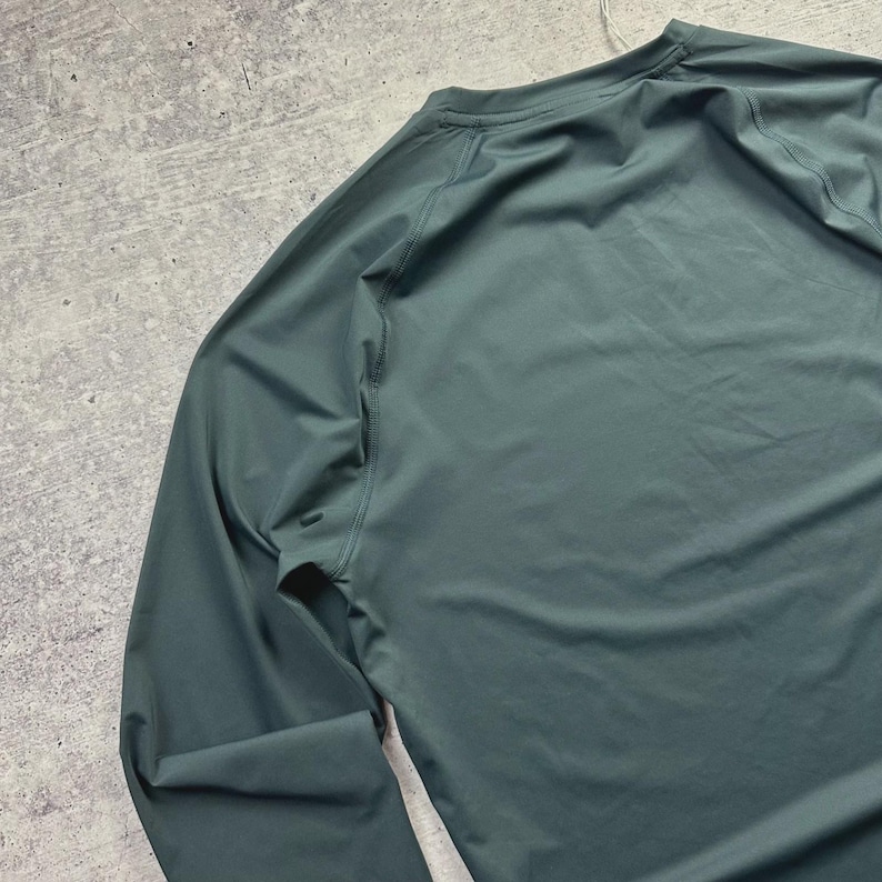May include: A long-sleeved, teal-colored athletic shirt. The shirt has a crew neck and raglan sleeves. The fabric appears to be a lightweight, moisture-wicking material, suitable for sports and exercise. The shirt is shown on a gray surface.