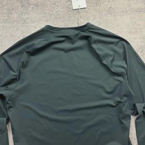May include: A long-sleeved, dark teal athletic shirt. The shirt has a crew neck and raglan sleeves. A white tag hangs from the collar. The fabric appears to be a lightweight, moisture-wicking material, suitable for sports or active wear.