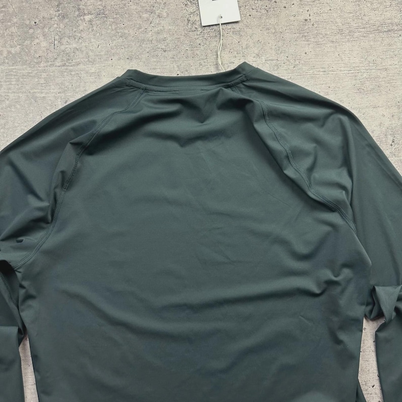 May include: A long-sleeved, dark teal athletic shirt. The shirt has a crew neck and raglan sleeves. A white tag hangs from the collar. The fabric appears to be a lightweight, moisture-wicking material, suitable for sports or active wear.