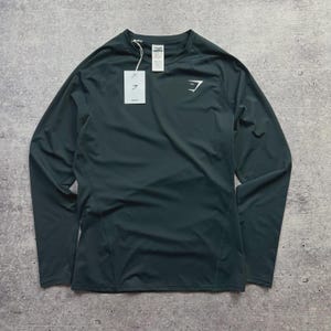 May include: Dark teal long-sleeve athletic shirt with a crew neck. The shirt features a white logo on the chest and a tag. The material appears to be a lightweight, performance fabric. The shirt is designed for athletic activities.