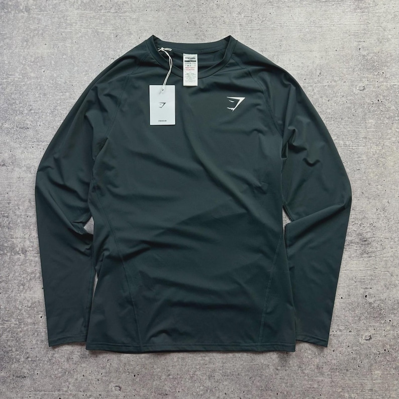 May include: Dark teal long-sleeve athletic shirt with a crew neck. The shirt features a white logo on the chest and a tag. The material appears to be a lightweight, performance fabric. The shirt is designed for athletic activities.
