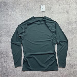 May include: A long-sleeved, dark teal athletic shirt. The shirt is made of a smooth, lightweight material and features a crew neck. A white tag hangs from the neckline. The shirt is displayed on a gray, textured surface.