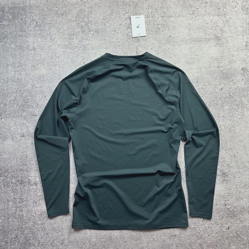 May include: A long-sleeved, dark teal athletic shirt. The shirt is made of a smooth, lightweight material and features a crew neck. A white tag hangs from the neckline. The shirt is displayed on a gray, textured surface.