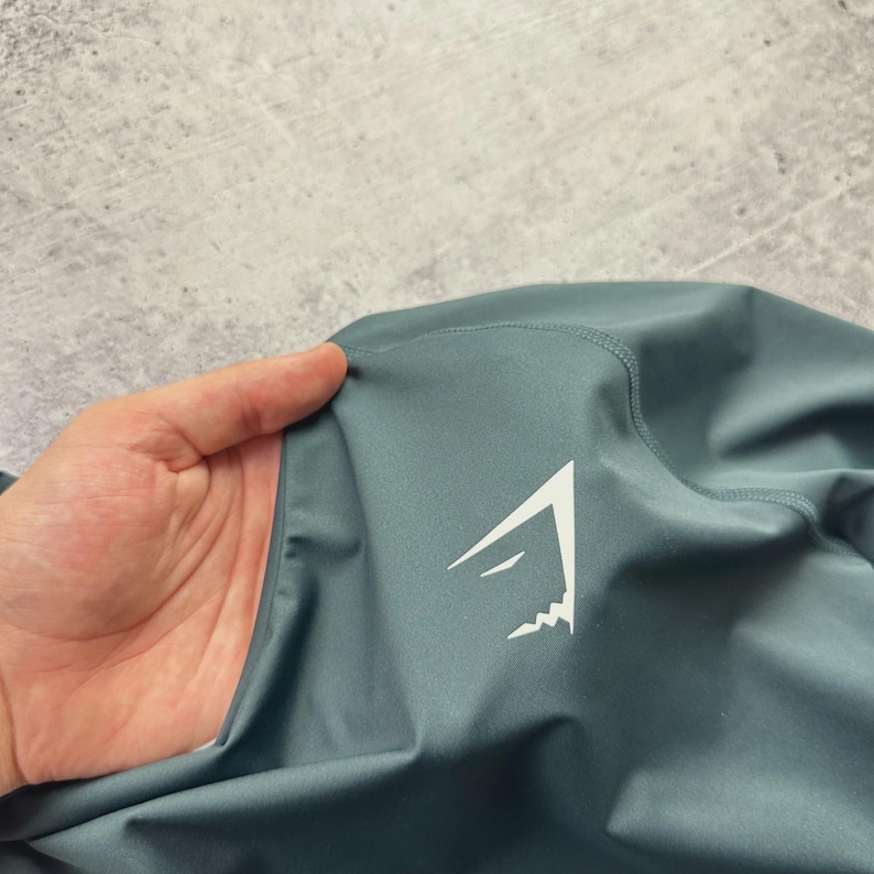 May include: Close-up of a teal athletic garment with a white logo. The logo is a stylized mountain peak design. The fabric appears smooth and slightly shiny, with visible seams.
