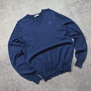 Puede incluir: Un suéter azul marino con cuello en V y mangas largas. El suéter tiene un dobladillo y puños acanalados, y un pequeño emblema bordado en el pecho. La prenda está hecha de un material de punto suave.