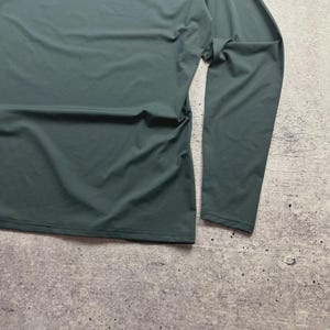 May include: A long-sleeved, dark teal shirt. The fabric is smooth and drapes softly. The shirt is laid flat on a textured, gray surface. The sleeves are long and end at the wrists.