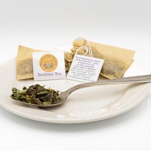 Puede incluir: Un plato de cerámica blanca con una cuchara llena de té de hojas sueltas y bolsitas de té. Las bolsitas de té están etiquetadas como "Bedtime Tea" y "Mountain Star Herbs". La imagen sugiere té de hierbas y relajación.