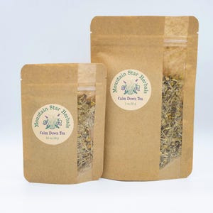 Puede incluir: Dos bolsas de papel marrón de té Calm Down de Mountain Star Herbals. Las bolsas tienen una ventana transparente que muestra la mezcla de té. Una bolsa está etiquetada 0,5 oz/14 g, y la otra 1 oz/28 g.