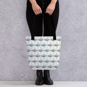 May include: A white tote bag with a geometric pattern of gray and blue triangles. The bag has black handles and is being held by a person.