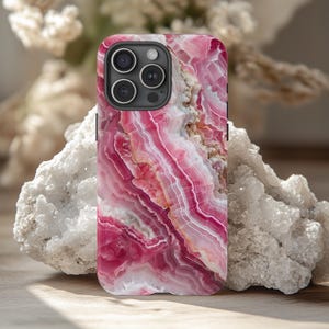 May include: A pink and white agate phone case with a camera lens cutout. The case has a swirling pattern of pink and white.