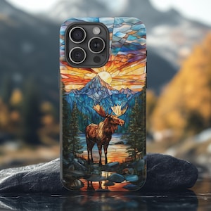 May include: A phone case with a stained-glass effect depicting a moose by a lake, with mountains and a sunset. The case has a black frame and a glossy finish. The design is a detailed, colourful illustration.
